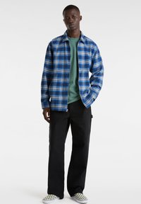 Blau-weißer Karo-Flanelljacke, geschichtet über einem grünen Rundhalsoberteil, kombiniert mit schwarzen Hosen und karierten Slip-On-Schuhe.