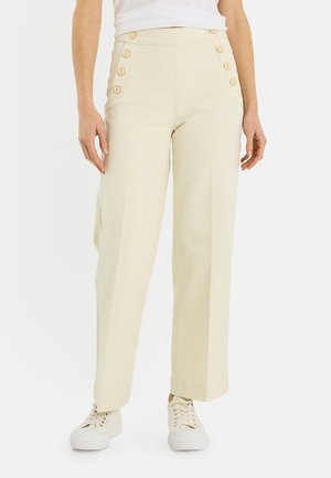 Femme portant un pantalon crème taille haute avec des boutons sur le côté et des baskets blanches, debout devant un fond blanc.