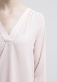 Ljusrosa blus med V-urringning. Tillverkad av ett mjukt tyg, med långa ärmar och subtila rynkor vid axlarna.