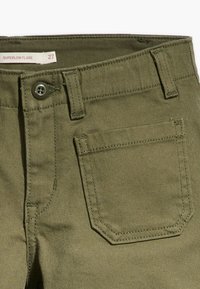 Pantalon flare très bas en tissu de coton vert olive. Comprend une fermeture à boutons, cinq poches et des détails de couture subtils.