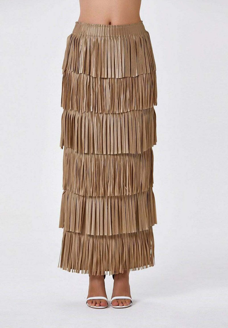 Jupe maxi à franges en brun clair, comportant plusieurs bandes de tissu superposées. Taille élastique et surface texturée. Portée avec des sandales blanches.