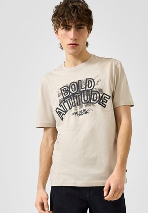 Junger Mann mit lockigem braunem Haar, der ein beiges T-Shirt mit dem Text "Bold Attitude" und dunkle Hose vor einem einfarbigen hellen Hintergrund trägt.
