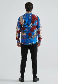Chemise à manches longues avec un fond bleu vif, ornée de divers motifs floraux et imprimés animaliers, et un col à boutons. Portée avec un pantalon noir.