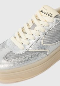 Sidabriniai ir pilki sportbačiai su kreminėmis raišteliais, tekstūruota oda ir tinkliniai akcentai, bei smėlio spalvos padas, su etiketėmis "Scotch & Soda" ant liežuvėlio.
