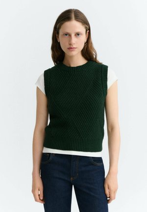 Jeune femme aux longs cheveux bruns porte un gilet sans manches tricoté vert foncé par-dessus une chemise blanche à manches courtes et un jean bleu foncé.