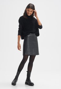 Pullover oversize nero in maglia con collo alto, abbinato a una gonna nera in pelle aderente con un design a patchwork, collant neri e stivali chunky.