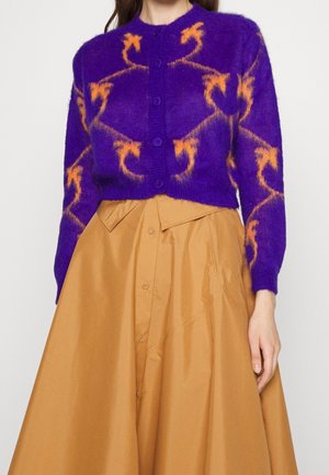 Paarse harige cardigan met oranje kettingachtig patroon, vooraan met knopen, gecombineerd met een hooggesneden beige rok met knopen aan de voorkant en brede plooien.
