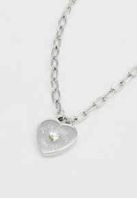 Silver heart-shaped necklace with embossed text "IN LOVE SINCE 1981," featuring a central clear gem and chain links.