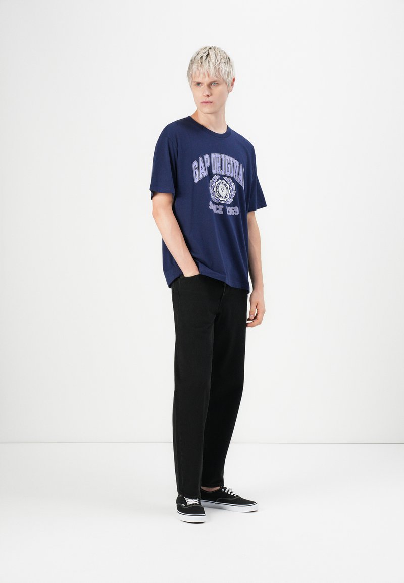 Camiseta de algodón azul marino con el logo de GAP en blanco y púrpura, combinada con pantalones negros y zapatillas negras, de pie contra un fondo blanco.