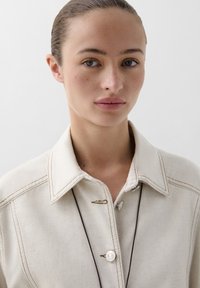 Beige collared shirt with visible stitching, a button closure, and a decorative hook detail. Smooth texture and neutral tone, minimal design.