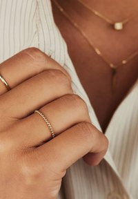 Anello placcato in oro con un design a perline su una mano. La mano è parzialmente in ombra, mostrando una consistenza della pelle liscia. Visibile una camicia a righe.