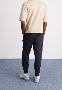 Camisa beige de manga corta con un corte holgado, combinada con pantalones cargo negros que tienen bolsillos laterales. Zapatillas de deporte grises con cámaras de aire visibles.