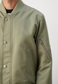 Man wearing a light olive green bomber jacket with snap buttons and a zippered pocket on the upper left sleeve against a plain background.