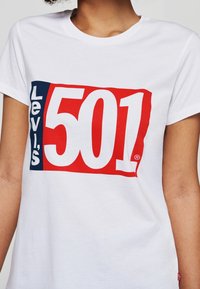Bílá bavlněná trička s velkým červeným a modrým potiskem, na kterém je výrazně napsáno "Levi's 501". Kulatý výstřih, krátké rukávy.