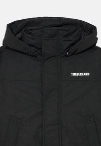 Svart vadderad jacka med hög krage, huva och framdragkedja. "TIMBERLAND"-logotyp i vitt på vänster bröst. Slät textur.