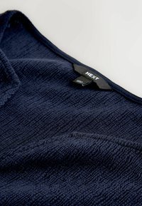 Marineblåt tekstureret stofbeklædning med en firkantet udskæring og en sort tøjetikette med teksten "NEXT" ved kraven.