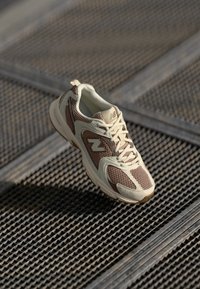 Beige and brown athletic shoe with mesh upper, cushioned sole, and lace-up design. Features logo accent and textured overlays.