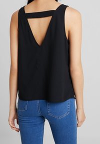 Femme portant un haut noir sans manches avec un dos en V profond et une sangle horizontale, associée à un jean en denim bleu, debout devant un fond uni.