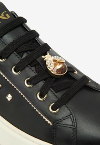 Sneaker en cuir noir avec des lacets noirs, des œillets dorés, des clous dorés et un charm décoratif en forme d'abeille en or et strass sur les lacets.