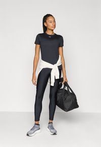 Μαύρο αθλητικό μπλουζάκι, εφαρμοστά μαύρα κολάν, κρεμ πουλόβερ δεμένο στη μέση, γκρι αθλητικά παπούτσια και μια μαύρη τσάντα ντούφλ.