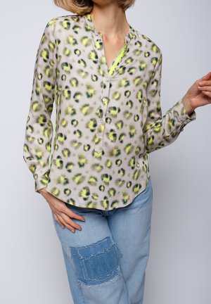 Femme portant un chemisier beige à manches longues avec un imprimé léopard jaune et noir, associé à un jean bleu clair avec une grande pièce en denim.