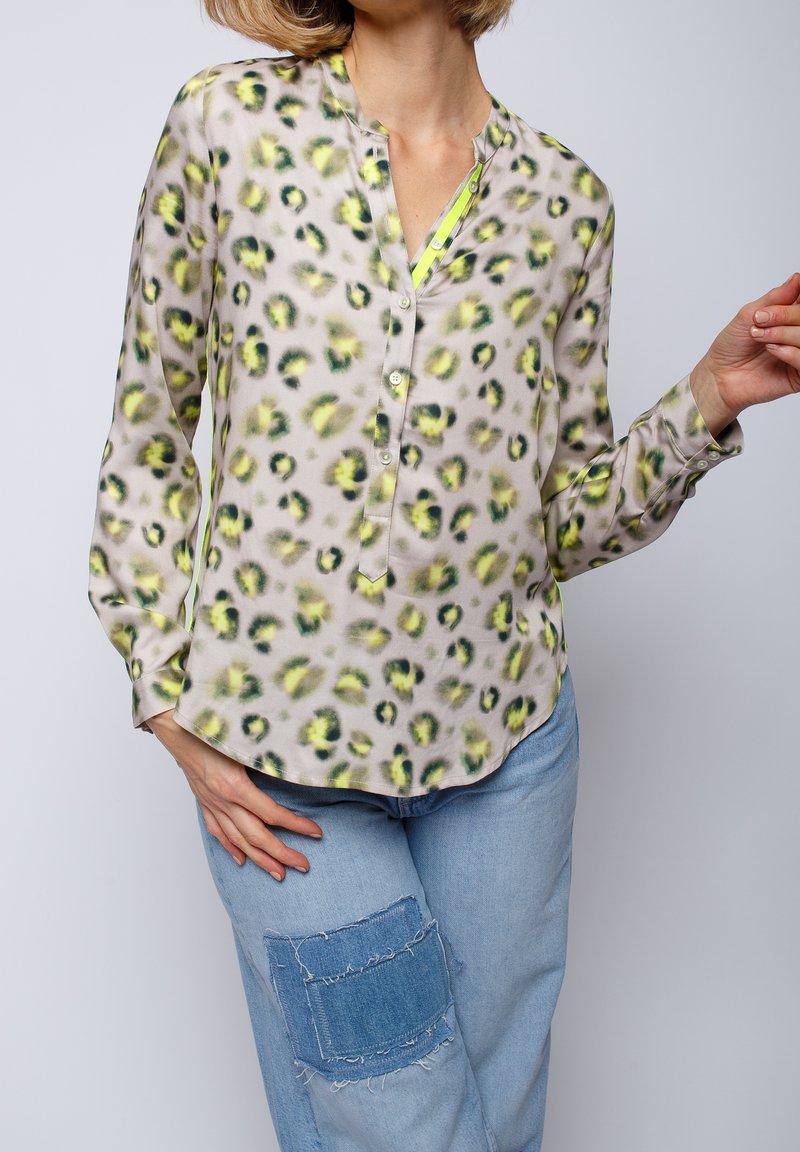 Femme portant un chemisier beige à manches longues avec un imprimé léopard jaune et noir, associé à un jean bleu clair avec une grande pièce en denim.