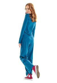 Mėlynos spalvos sportinis kostiumas, laisvas kirpimas, ilgos rankovės ir siaurėjantys kelnių modeliai. Derinamas su raudonomis aukštakulnėmis sportinėmis bateliais. Minkšta medžiagos struktūra, minimalus dizainas.