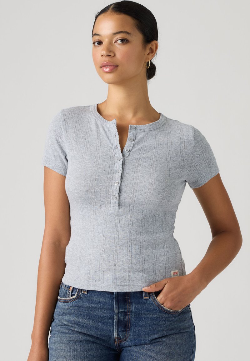 Femme aux cheveux foncés en chignon bas, portant une chemise côtelée gris clair à manches courtes boutonnée et un jean bleu, une main dans la poche.