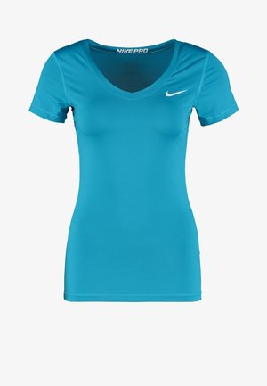 Femme portant un t-shirt de sport bleu vif, un short bleu foncé avec des accents bleu clair, et des baskets blanches avec des lacets jaune néon, debout droite.