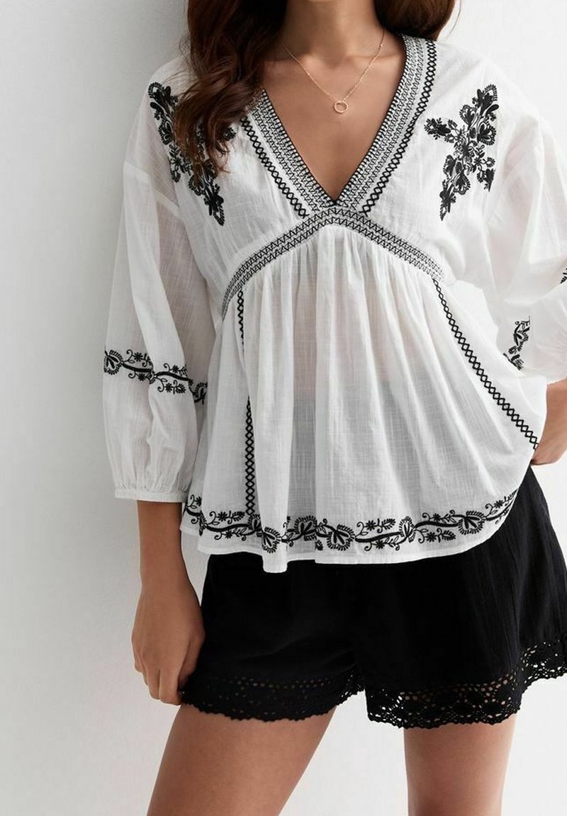 Femme portant un chemisier blanc avec broderie florale noire et un short rehaussé de dentelle noire, debout avec une main dans la poche.