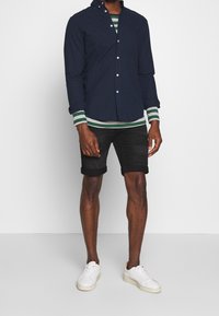 Merijälle painettu nappipaita vihreän raidallisen neuleen päällä, yhdistetty mustiin farkkushortseihin ja valkoisiin lenkkareihin. Shortseissa on käänteiset lahkeet.