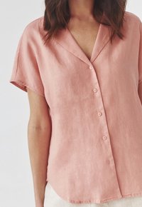Blusa di lino rosa con scollo a V, maniche corte e cinque bottoni sul davanti. Tessuto liscio, vestibilità rilassata e orlo curvo.