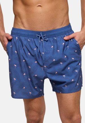 Hombre con pantalones cortos de baño azules con pequeños estampados de flamencos rosa y hojas, manos en los bolsillos, sobre un fondo liso.