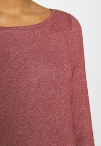 Gros plan sur une personne portant un haut en maille à manches longues, doux, de couleur rose rougeâtre, avec un col rond, sur un fond uni.