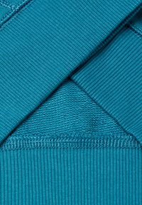 Tessuto teal con texture rigate e lisce, caratterizzato da un taglio trasversale di due strati. Cuciture visibili lungo i bordi.