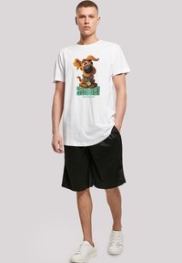 Hvit bomull t-skjorte med et tegneseriehund-graphic i brunt, svart og beige, sammen med teksten "SCOOB!" i grønt. Bært sammen med svarte shorts og hvite sneakers.