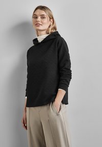 Pullover nero con cappuccio e motivo a diamante testurizzato, dotato di maniche lunghe e vestibilità comoda. Abbinate a pantaloni beige chiaro plissettati.