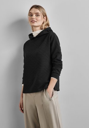 Schwarzer Kapuzenpullover mit strukturiertem Rautenmuster, der lange Ärmel und eine lockere Passform hat. Kombiniert mit hellbeigen, plissierten Hosen.