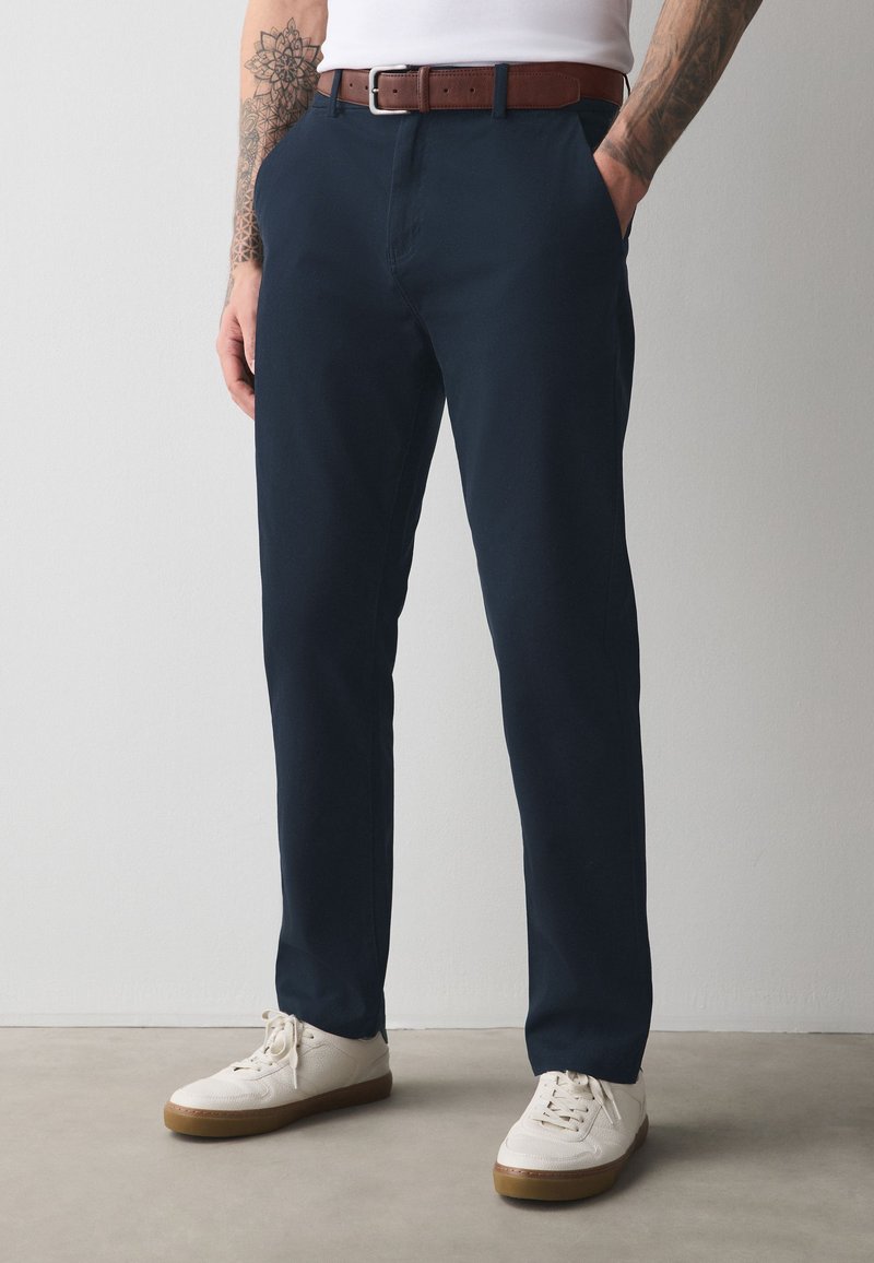 Mann trägt marineblaue Hose, weiße Sneaker mit Gummisohlen, braunen Ledergürtel und weißes Hemd, steht mit den Händen in den Taschen.