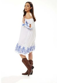 White off-shoulder dress with blue floral embroidery, ruffled hem, paired with brown knee-high cowboy boots featuring stitch detailing.
