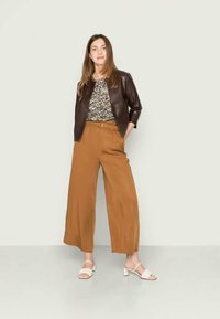 Casaco de pele castanha sobre uma blusa floral, combinado com calças largas em tom bege e sandálias brancas. Texturas suaves e cores contrastantes.