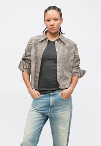 Osoba z zaplecionymi włosami ubrana w koszulę w paski z guzikami na czarnym topie oraz jasnoniebieskie, przetarte dżinsy, stoi z rękami w kieszeniach.