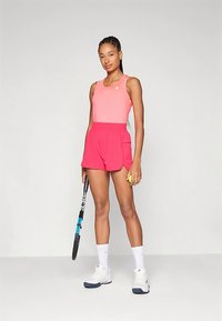 Γυναίκα με ροζ αμάνικο τοπ και σορτς, κρατώντας μια ρακέτα τένις και μια μπάλα, στέκεται μπροστά σε έναν απλό ανοιχτό φόντο.