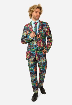 Homme portant un costume coloré et une cravate assortie arborant des personnages de dessins animés et des motifs psychédéliques, avec des chaussures noires sur fond blanc.