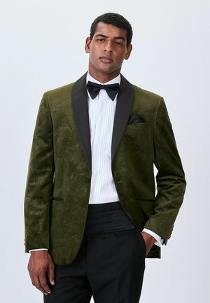 Mees tumesinise mustriga sametist smokingutagi mustade krae, valge volditud särk, must lips ja mustad püksid, lihtsa tausta ees.