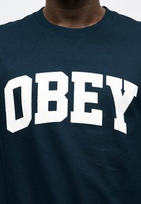 Σκούρο μπλε μπλουζάκι με στρογγυλή λαιμόκοψη και μεγάλα λευκά έντονα γράμματα που σχηματίζουν τη λέξη «OBEY» στο στήθος, φορεμένο από ένα άτομο.