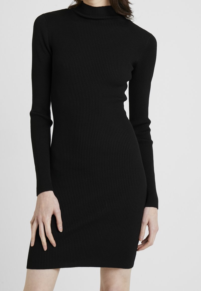 Robe noire ajustée en maille côtelée pour femmes, à manches longues et col montant, présentée de la nuque jusqu'à mi-cuisse sur un fond uni.