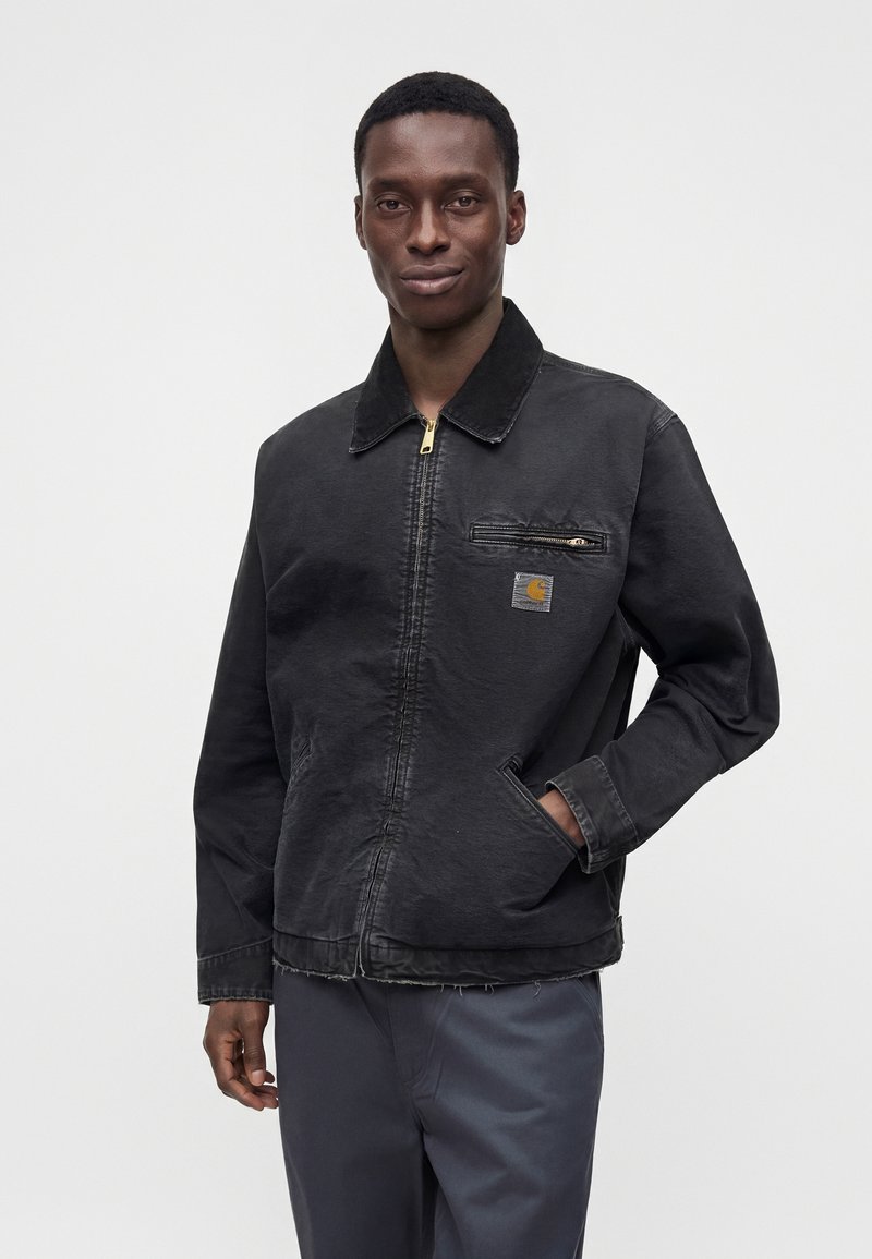 Mann trägt eine schwarze Jacke mit Reißverschluss und Brusttasche mit Reißverschluss und Logo-Aufnäher, steht mit einer Hand in seiner Seitentasche.