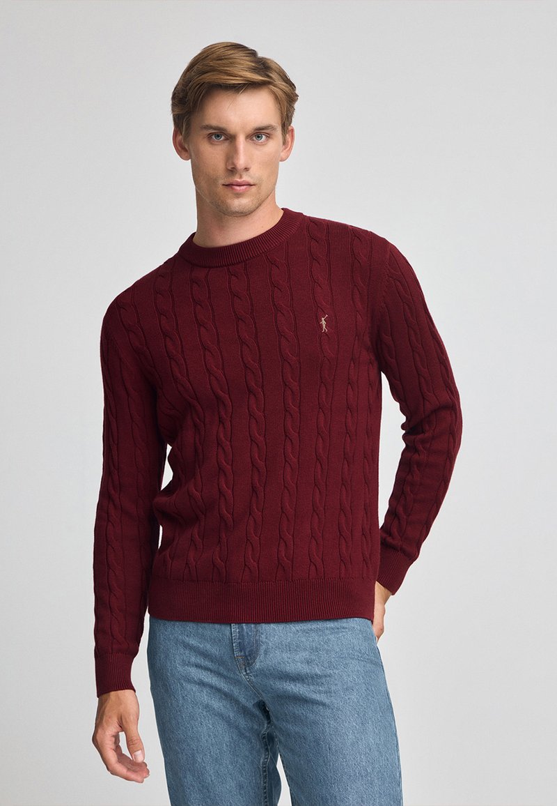 Burgundy kabelstickad tröja med rund halsringning och ribbade ärmslut, med en subtil logotyp broderad på bröstet.