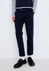 Marina bomullbyxor med slim fit, tillsammans med ett svart bälte med cirkulära hål. Bärs med svarta skor och vita strumpor. Randig långärmad topp.